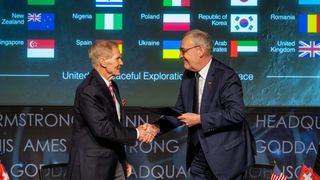 two white men in dark suits shake hands in front of a backdrop of many flags.